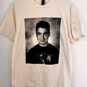 May include: Cream-colored t-shirt featuring a black and white portrait of a person. The portrait is centered on the shirt. The person in the portrait is smiling. The shirt has short sleeves and a crew neck.