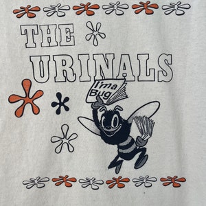May include: A white t-shirt with a black and white graphic of a bee holding a newspaper that says "I'ma Bug". The text "THE URINALS" is printed in black above the bee. The shirt is decorated with orange and white flower-shaped designs.