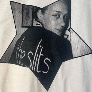 以下が含まれることがあります： 黒の星形のデザインが施された白いTシャツ。星には、白黒の女性の画像と白い文字で「the slits」と書かれています。