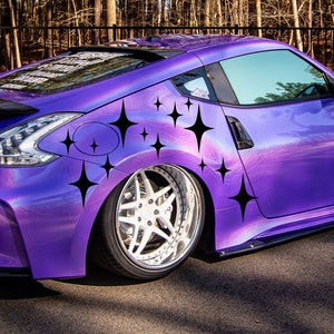 Puede incluir: Un Nissan 370Z morado con calcomanías de estrellas negras en el lateral. El coche está estacionado en un camino pavimentado frente a un bosque.