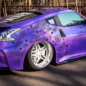 Puede incluir: Un coche morado con un envoltorio de patrón paisley. El coche está aparcado en una superficie pavimentada con un bosque en el fondo.