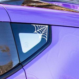May include: A purple car with a black spoiler and a white spiderweb decal on the rear window. The car has a black door handle and white wheels.