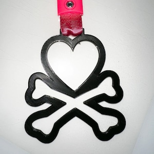 Op de afbeelding: Een zwarte hart- en kruisbottenhanger met een roze band. De hanger heeft een hartvorm in het midden en is verbonden met een roze band met een roze plastic stuk en twee zilveren klinknagels.