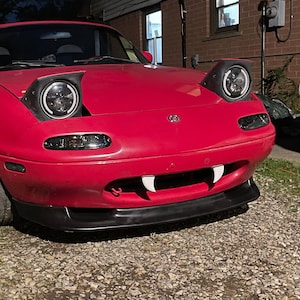 May include: A red Mazda Miata convertible with black headlights and a black front bumper. The car has a white fang-like design on the front bumper.