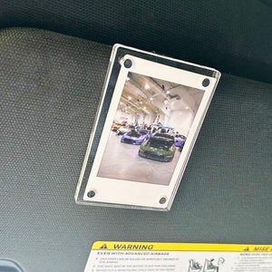 May include: A clear plastic photo frame with a picture of a car show inside. The frame is attached to a car's sun visor. The warning label on the visor reads "Warning Even with Advanced Airbags".