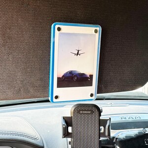 May include: A black phone holder with a textured surface is mounted on a car's air vent. A blue and white photo frame with a picture of an airplane flying over a car is attached to the sun visor.