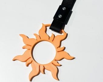 May include: A peach-colored sun-shaped keychain with a black adjustable strap. The sun has a circular center and 12 points radiating outward.