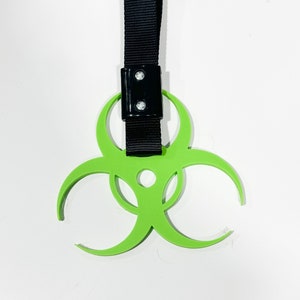 May include: A neon green biohazard symbol with a black strap and a black rectangular connector. The symbol is a three-lobed design, and the strap is made of a woven material.