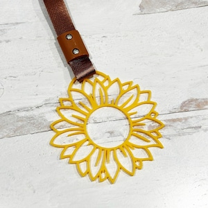 Può includere: Un portachiavi giallo a forma di girasole con un cinturino marrone. Il cinturino ha due bottoni a pressione metallici.