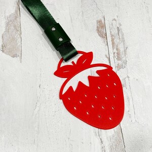Può includere: Un portachiavi a forma di fragola rossa con un cinturino verde. La fragola ha un centro bianco e un gambo verde.