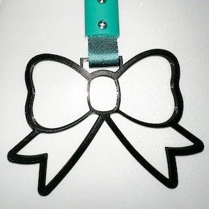 Può includere: Un portachiavi in ​​plastica nera a forma di fiocco con un laccio turchese e un fermaglio in plastica turchese. Il fermaglio ha due viti argentate.