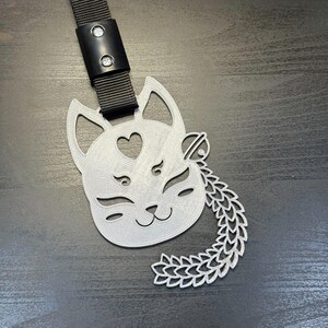 Può includere: Accessorio a forma di gatto color argento con cinturino nero. Il volto del gatto presenta un dettaglio a forma di cuore e una coda decorativa. Il cinturino è fissato con una fibbia nera.