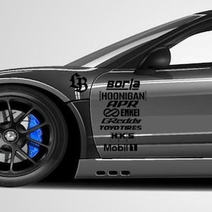 May include: A grey sports car with black wheels and blue brake calipers. The car features sponsor decals, including "BORLA" and "HOONIGAN". The car is set against a plain white background.