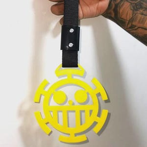 May include: A yellow plastic handle featuring the circular Heart Pirates emblem with a wide, toothy grin and two round eyes. The emblem is attached to a black nylon strap secured by a black rectangular connector with two silver-colored metal rivets.
