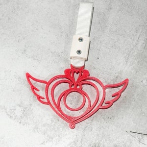 Può includere: Un ciondolo di plastica rosso a forma di cuore con le ali. Il ciondolo è attaccato a un cinturino bianco con una fibbia di plastica bianca.
