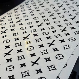 Puede incluir: Hojas de pegatinas blancas con un patr&oacute;n negro repetido de flores de cuatro p&eacute;talos, cruces y c&iacute;rculos entrelazados. Las pegatinas est&aacute;n dispuestas en filas y columnas ordenadas, creando un dise&ntilde;o visualmente atractivo.