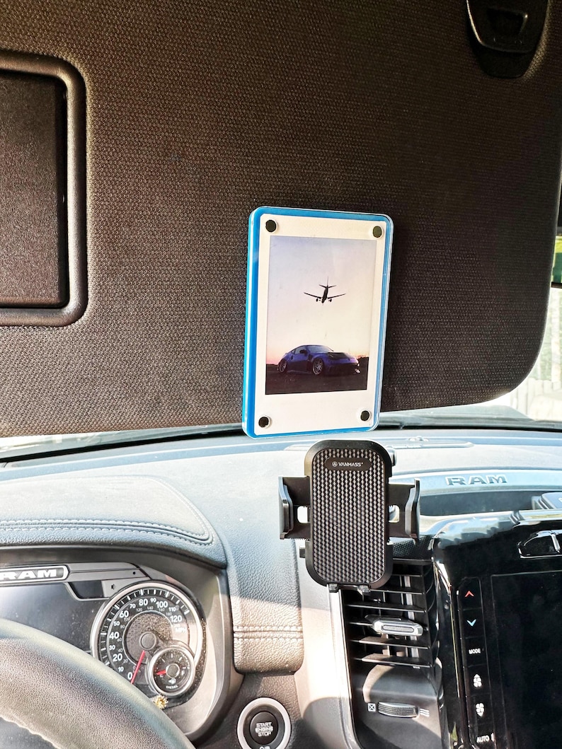 May include: A black phone holder with a textured surface is mounted on a car's air vent. A blue and white photo frame with a picture of an airplane flying over a car is attached to the sun visor.