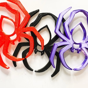 May include: Three 3D-printed spider decorations in red, black, and purple. The spiders have a stylized design with eight legs and a circular body. The decorations are likely for Halloween or a similar themed event.