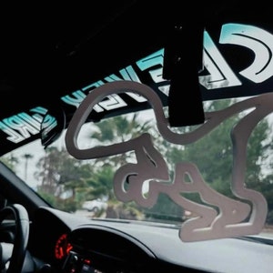 May include: White car air freshener shaped like a dinosaur hanging from the rearview mirror of a car. The car is parked and the trees outside are visible through the windshield.