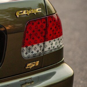Puede incluir: Coche verde oliva con emblemas dorados "CIVIC" y "SI". El piloto trasero presenta un dise&ntilde;o rojo y blanco con un patr&oacute;n de cruces y un logotipo repetido. El parachoques del coche refleja el entorno.