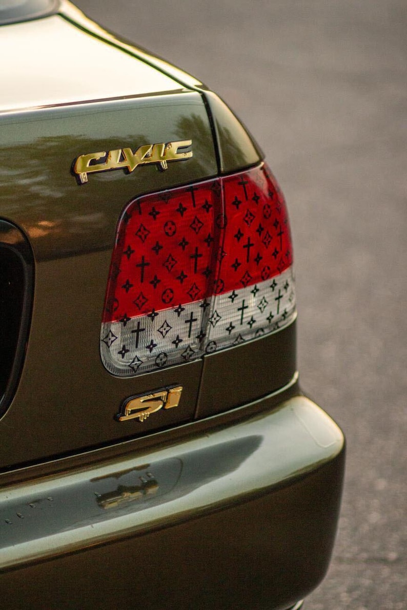 Puede incluir: Coche verde oliva con emblemas dorados "CIVIC" y "SI". El piloto trasero presenta un dise&ntilde;o rojo y blanco con un patr&oacute;n de cruces y un logotipo repetido. El parachoques del coche refleja el entorno.