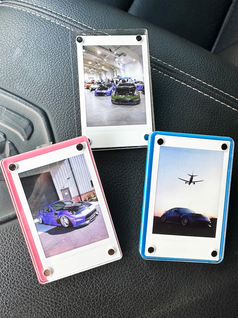 May include: Three clear plastic photo frames with different colored borders. The frames are holding instant photos. The first frame is clear with a white border and shows a photo of a green car in a car show. The second frame has a pink border and shows a photo of a purple car. The third frame has a blue border and shows a photo of a blue car with a plane flying overhead.