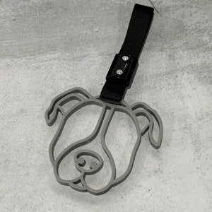 May include: A gray plastic dog head keychain with a black strap and a metal buckle. The dog head is a simple outline of a dog's face with ears, eyes, and a nose.