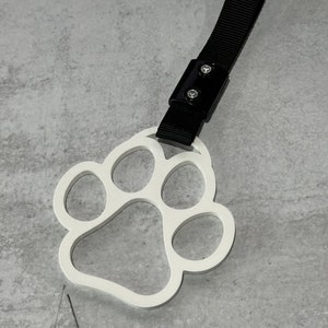 May include: White paw print shaped keychain with a black adjustable strap. The strap has a black plastic buckle with two silver screws.