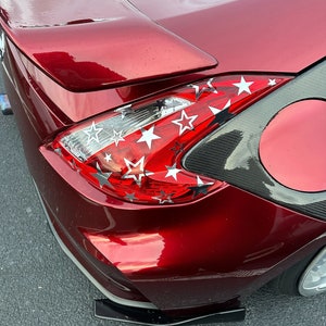 May include: A deep red car rear section featuring a prominent spoiler and a taillight assembly decorated with numerous silver and white star-shaped decals. The vehicle body includes a carbon fiber panel near the fuel cap and a black lower diffuser.