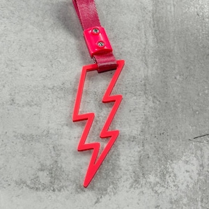 May include: A pink lightning bolt shaped keychain with a pink strap. The strap has two silver rivets.