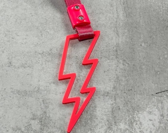 May include: A pink lightning bolt shaped keychain with a pink strap. The strap has two silver rivets.