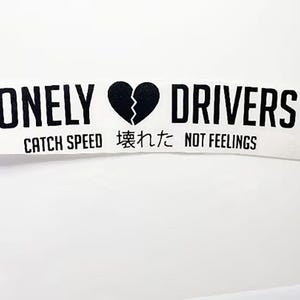May include: White sticker with black text and a broken heart graphic. The text reads "LONELY DRIVERS" with a broken heart symbol, and below it, "CATCH SPEED 壊れた NOT FEELINGS".