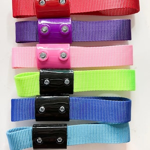 Könnte beinhalten: Eine Sammlung von sechs farbigen Nylonbändern mit Kunststoff- und Metallbeschlägen. Die Bänder sind in Rot, Lila, Pink, Neongrün, Blau und Hellblau gehalten. Jedes Band hat ein rechteckiges Kunststoffteil mit zwei Schrauben.