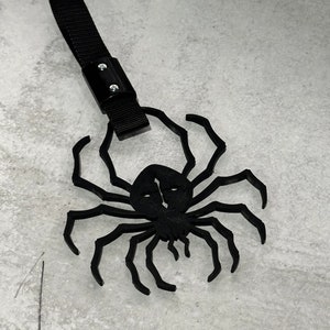 Könnte beinhalten: Schwarzes spinnenförmiges Accessoire mit schwarzem Riemen und Metallnieten. Die Spinne hat mehrere Beine und ein detailliertes Gesicht. Bei diesem Artikel handelt es sich wahrscheinlich um ein Dekorationsobjekt oder ein Halloween-Accessoire.