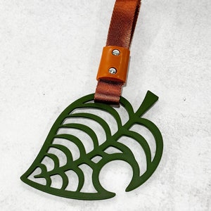 May include: A green leaf-shaped keychain with a brown strap and a brown plastic buckle. The leaf has a cut-out design.