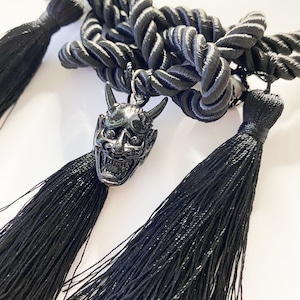 Op de afbeelding: Zwarte touwketting met drie zwarte kwastjes en een zilveren oni-masker hanger.