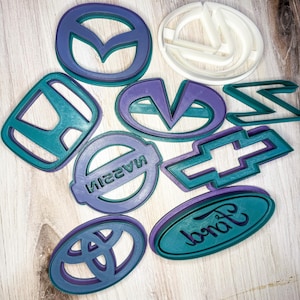 May include: A collection of car logo cookie cutters in teal and purple. The logos include Mazda, Nissan, Toyota, Ford, and Chevrolet. The Ford logo is an oval shape with the word "Ford" in cursive. The cookie cutters are on a light wood surface.