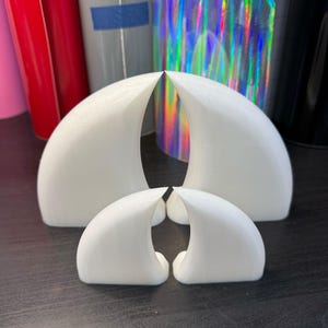 Puede incluir: Cuatro objetos blancos en forma de cuerno curvado, posiblemente para manualidades o decoración, están dispuestos sobre una superficie oscura. Los objetos varían en tamaño, con dos piezas más grandes y dos más pequeñas. Hay rollos de vinilo de colores en el fondo.