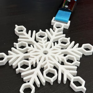 May include: A white, six-pointed snowflake ornament featuring geometric hexagonal cutouts at the ends of each branch. The snowflake is attached to a bright blue fabric strap secured by a polished silver-colored metal clamp with two visible screws.