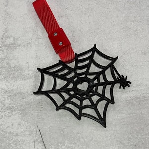 Op de afbeelding: Een zwarte spinnenweb-vormige ornament met een kleine spin aan de zijkant. Het ornament heeft een rode band die aan de bovenkant is bevestigd.