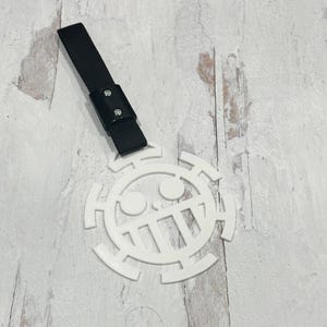 Puede incluir: Emblema circular blanco con un diseño de calavera y huesos cruzados, sujeto a una correa negra con una hebilla. El emblema tiene una calavera estilizada con un diseño de huesos cruzados. La correa es tejida.