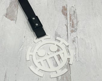 May include: White, circular emblem with a skull and crossbones design, attached to a black strap with a buckle. The emblem has a stylized skull with a crossbones design. The strap is made of a woven material.