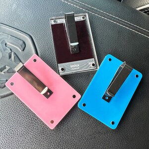 May include: Three plastic photo card holders with metal clips. The holders are pink, blue, and black. The black holder has the text "instax" and "FUJIFILM" printed on it.