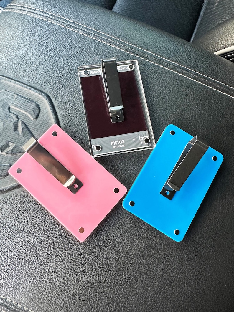 May include: Three plastic photo card holders with metal clips. The holders are pink, blue, and black. The black holder has the text "instax" and "FUJIFILM" printed on it.