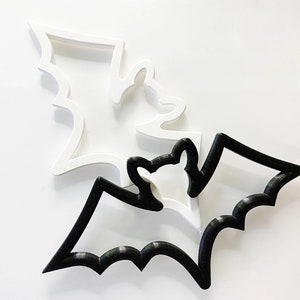 May include: Two bat-shaped cookie cutters, one white and one black. The black cutter is on top of the white cutter.