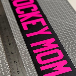 May include: Black rectangular patch with the words "HOCKEY MOM" in bright pink block letters. The patch is on a grey cutting mat with a metal ruler to the left.