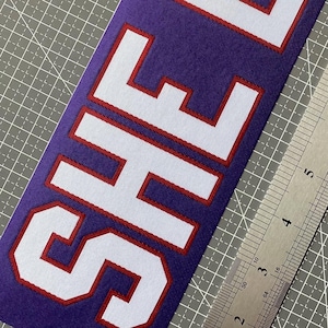 May include: White fabric patch with the word "SHEFF" in red outlined letters on a purple background.