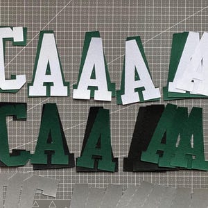 May include: Felt letters in green, black, and white spelling "CAA" and "M". Some letters have a white outline. The letters are arranged on a gray cutting mat, showing a craft project in progress.