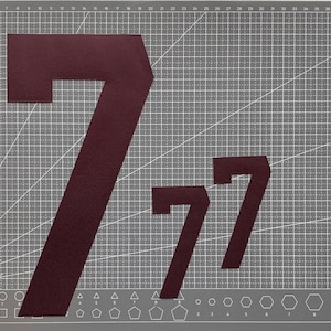 May include: A gray cutting mat with a grid pattern and geometric shapes along the bottom edge. Three burgundy number 7s are displayed on the mat, varying in size. The mat has the text "deli" and "No. 7840" in the bottom right corner.