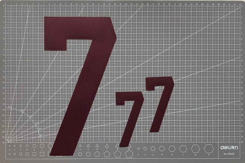 May include: A gray cutting mat with a grid pattern and geometric shapes along the bottom edge. Three burgundy number 7s are displayed on the mat, varying in size. The mat has the text "deli" and "No. 7840" in the bottom right corner.